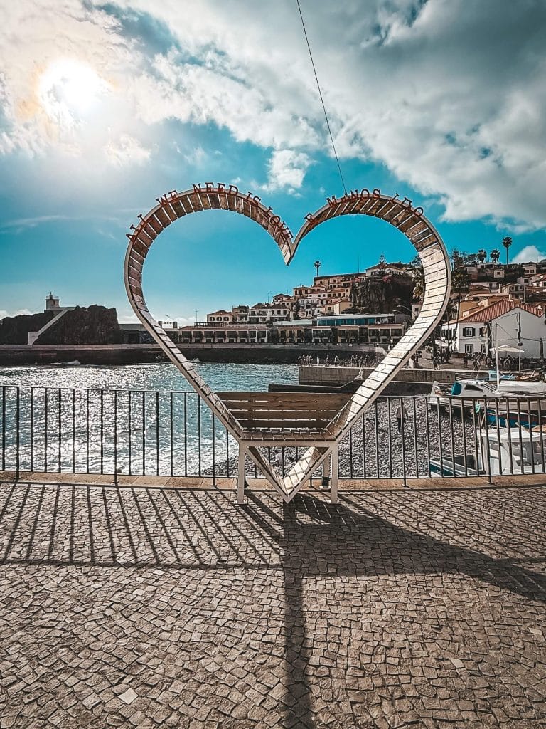 A heart-shaped metal sculpture with the words “All we need is love” on stone paving above the sea, overlooking the bay and the buildings of Câmara de Lobos, against the sun.