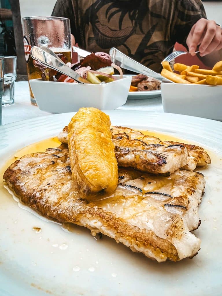 A plate of grilled fish fillet topped with a piece of fried banana in butter sauce; in the background a second plate with espetada, chips and a Coral beer.