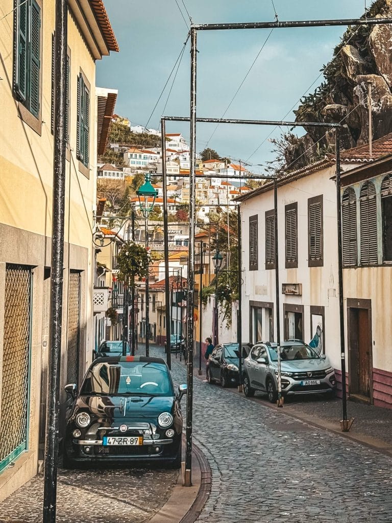 A narrow cobbled street with colourful buildings on both sides, a Fiat 500 parked at the kerb, cables strung between the houses and buildings on the hillside behind