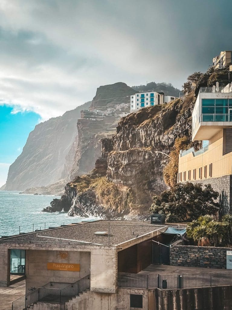 Rocky cliffs with buildings (including Praia do Vigário and a modern hotel) built into the cliff edge, the ocean on the left and the massive Cabo Girão fading into the mist behind.