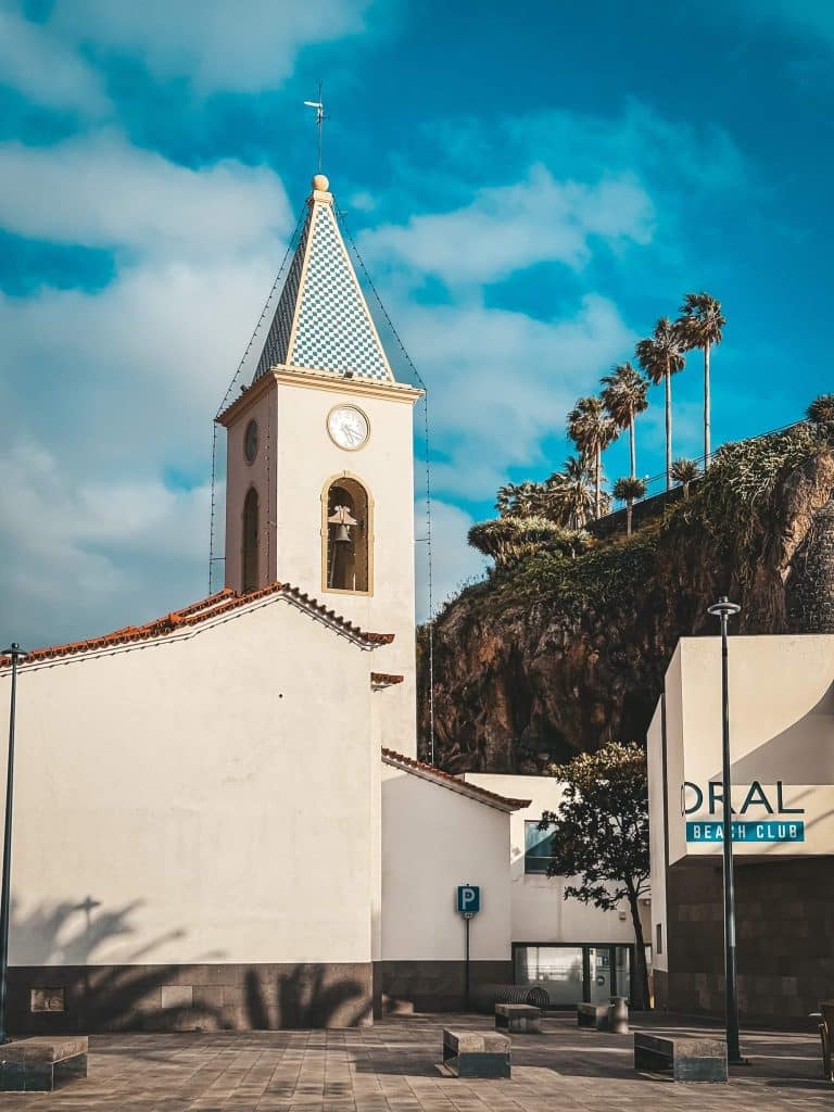 A white church with a tower topped by a green-and-white chequerboard pattern, with a clock, palm trees and the modern Coral Beach Club building on the right, against a rocky cliff.