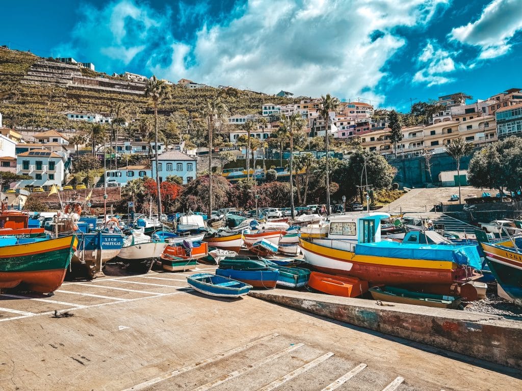 A wide shot of the harbour of Câmara de Lobos with dozens of colourful fishing boats on a concrete quay, terraced vineyards on a steep slope and colourful houses piled up the hill under a dramatic cloudy sky.