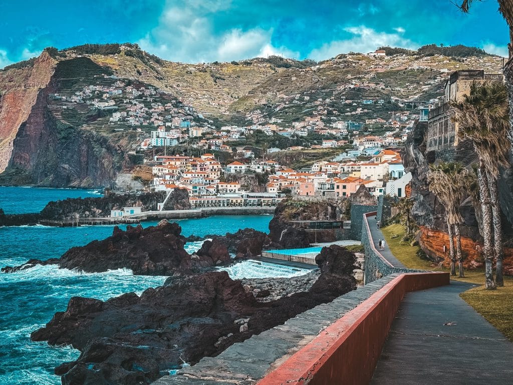 View from the seafront promenade with a red railing over dark lava rocks rising from the turquoise ocean, natural bathing pools, and the colourful buildings of Câmara de Lobos stacked up the slope below a massive cliff.