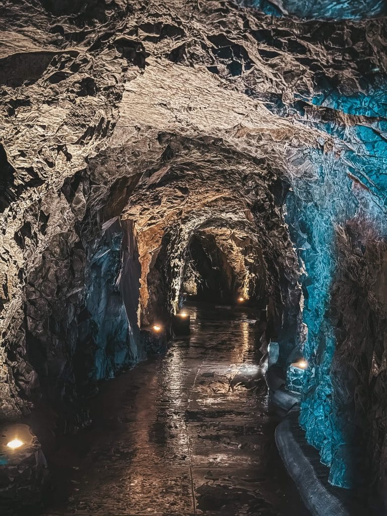 Inside a rock tunnel lit by warm and blue lamps, with a wet path leading in and a levada running along the wall.