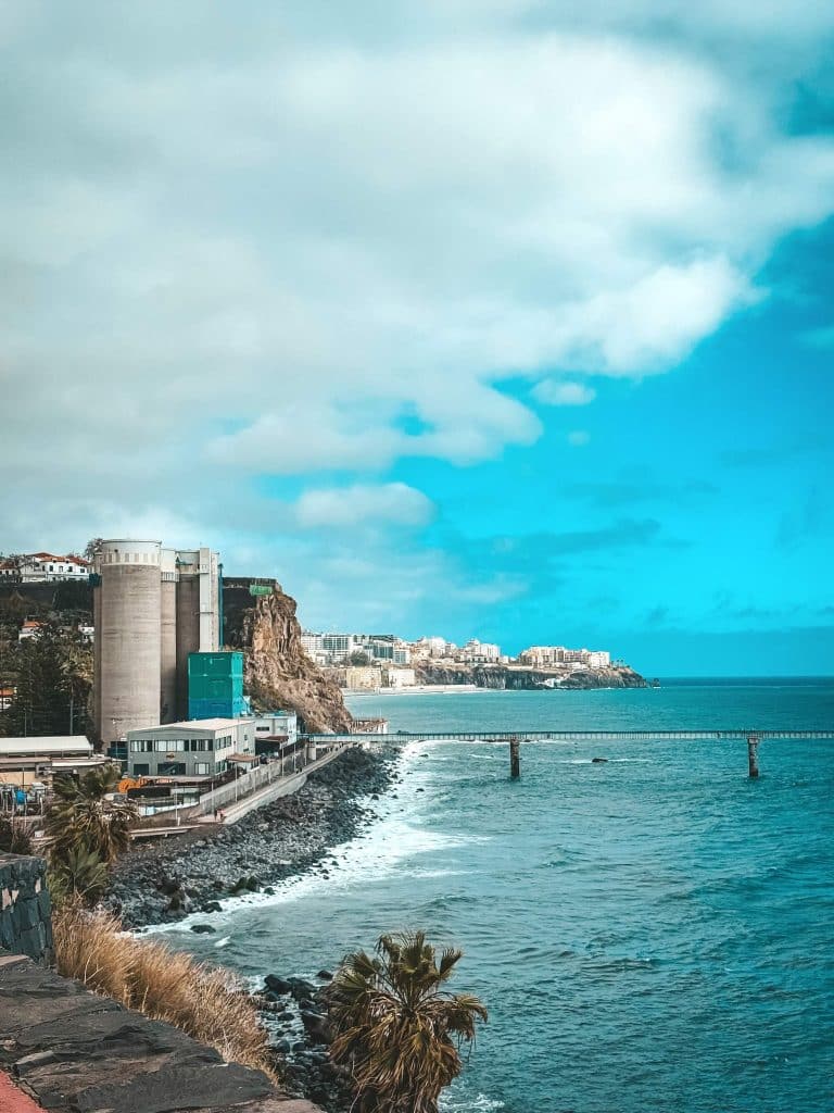 The waterfront with concrete silos and industrial buildings on the left, a stony shore and a bridge on piles over the turquoise ocean, with the city on the cliff behind.