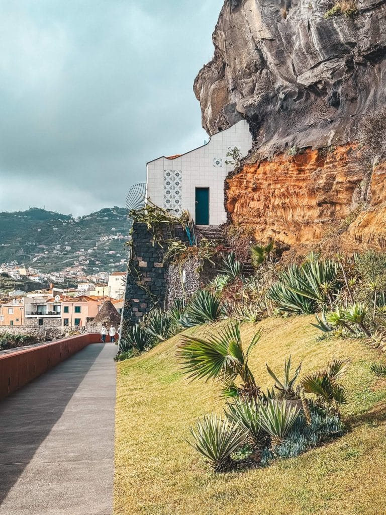 A small white building with decorative tiles and a turquoise door, built into an orange-brown cliff, with a lawn and palm trees in the foreground and the town behind.