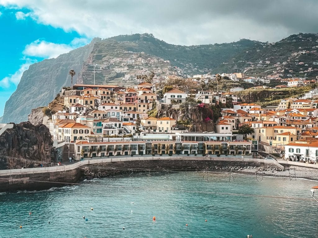 A wide panorama of the bay of Câmara de Lobos with turquoise water, a pebble beach, a row of waterfront buildings with restaurants and colourful houses on a steep slope, with a huge cliff behind.