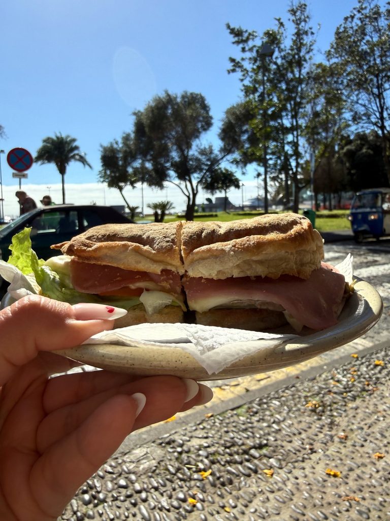 A hand holding a plate with a sandwich in a round bread roll with meat and lettuce, palms and trees behind