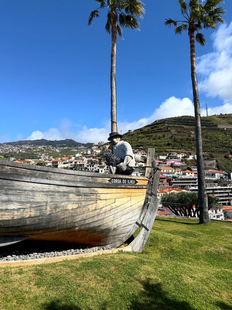 A wooden fishing boat with the name “Coroa do Ilhéu” set on a lawn as a monument, a figure of a fisherman in a hat on the bow, palm trees and houses of Câmara de Lobos behind