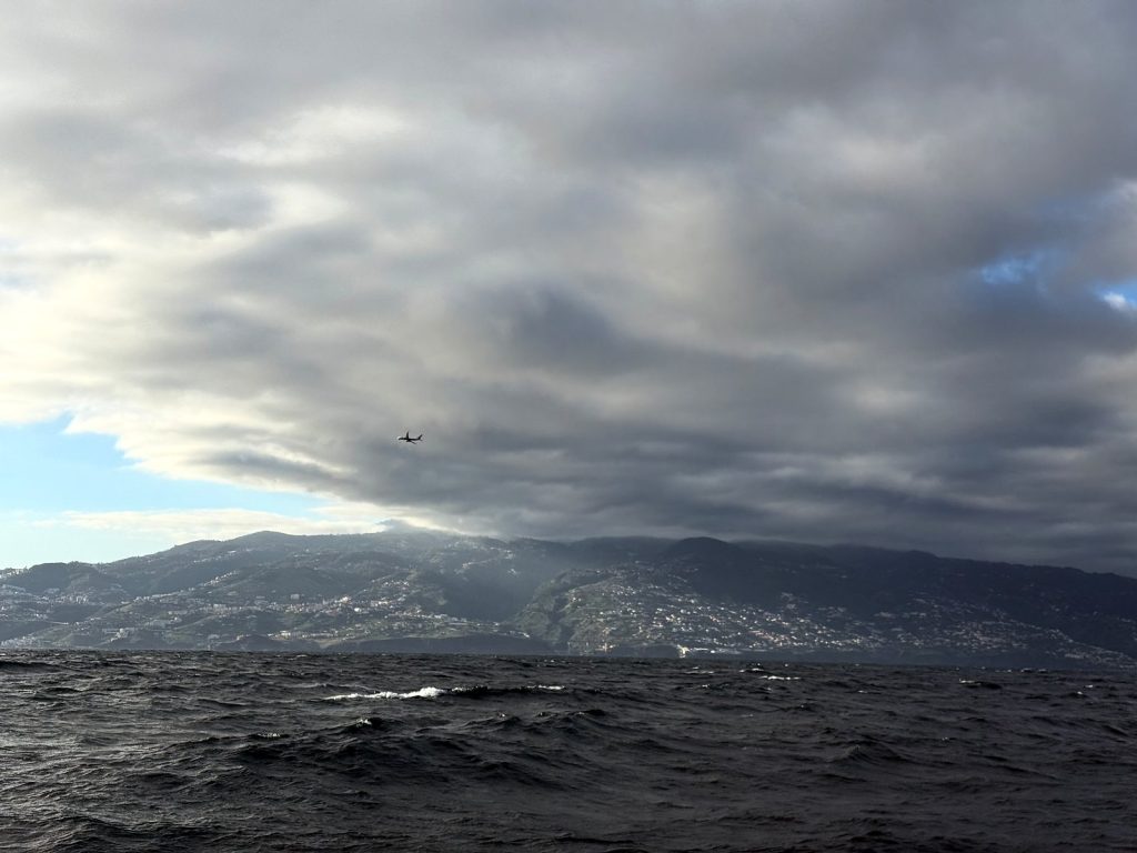 A thick layer of grey cloud over the slopes of Madeira’s coast seen from the ocean, with a plane in the sky