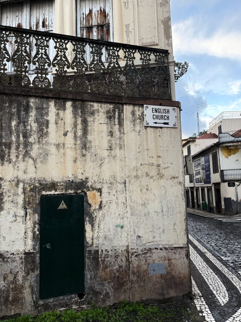 The peeling wall of an old town house with a wrought-iron balcony and green doors, a white English Church sign on the corner, a cobbled lane beside it