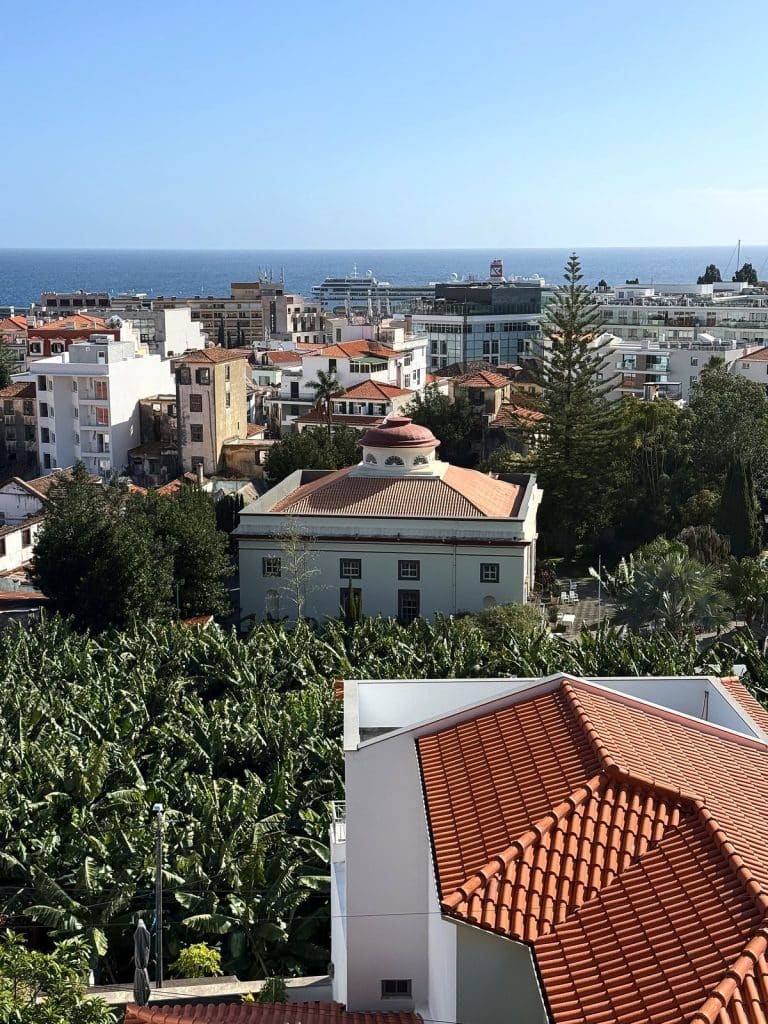 A white church with a small dome seen from above, a banana plantation in front, town houses of central Funchal and the ocean behind