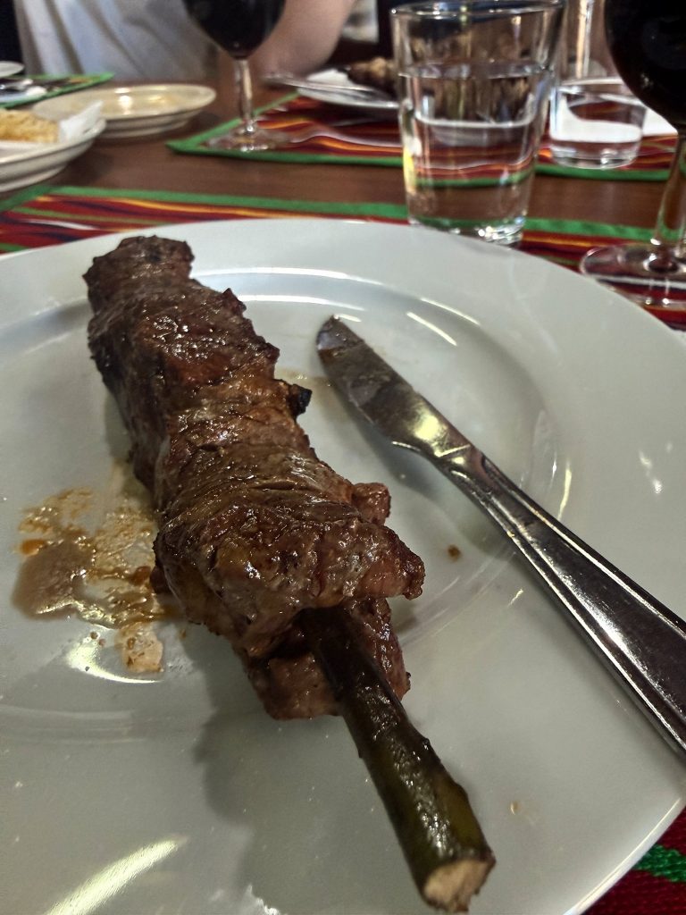 Espetada on a wooden skewer on a white plate, with a knife beside it and glasses of red wine and a colourful striped tablecloth behind
