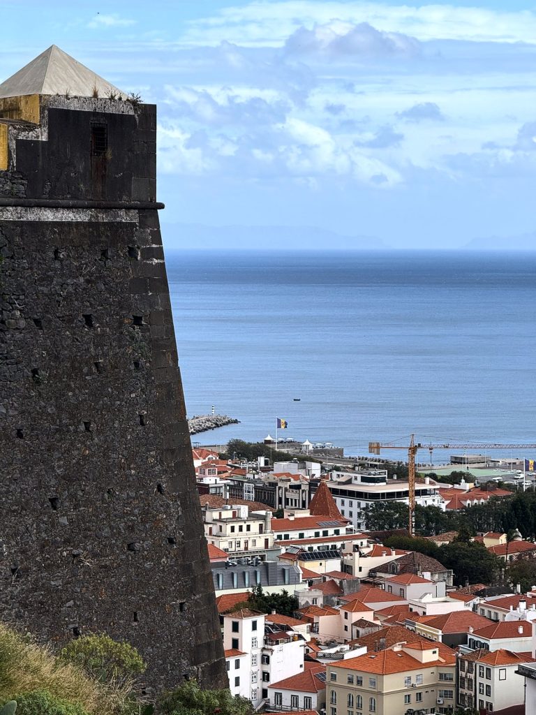 A dark stone watchtower of the fort with a pyramid roof, next to it a view down over Funchal’s red roofs, the harbour and the ocean