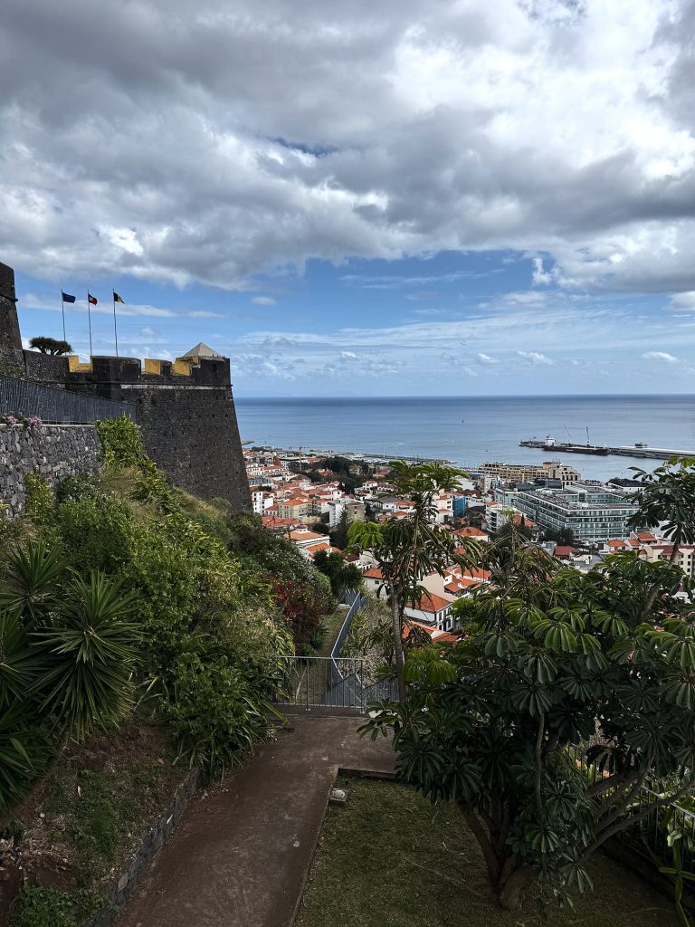 A path through greenery below the walls of Fortaleza do Pico, three flags on the walls and a corner watchtower with yellow battlements, Funchal’s rooftops, the harbour and the ocean behind