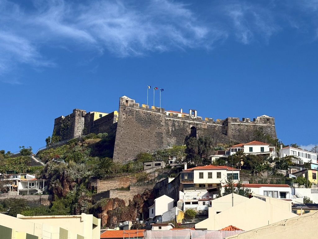 Fortaleza do Pico – the dark basalt walls of the fortress with three flags on top of the hill, houses with red roofs below, a blue sky with wispy clouds above