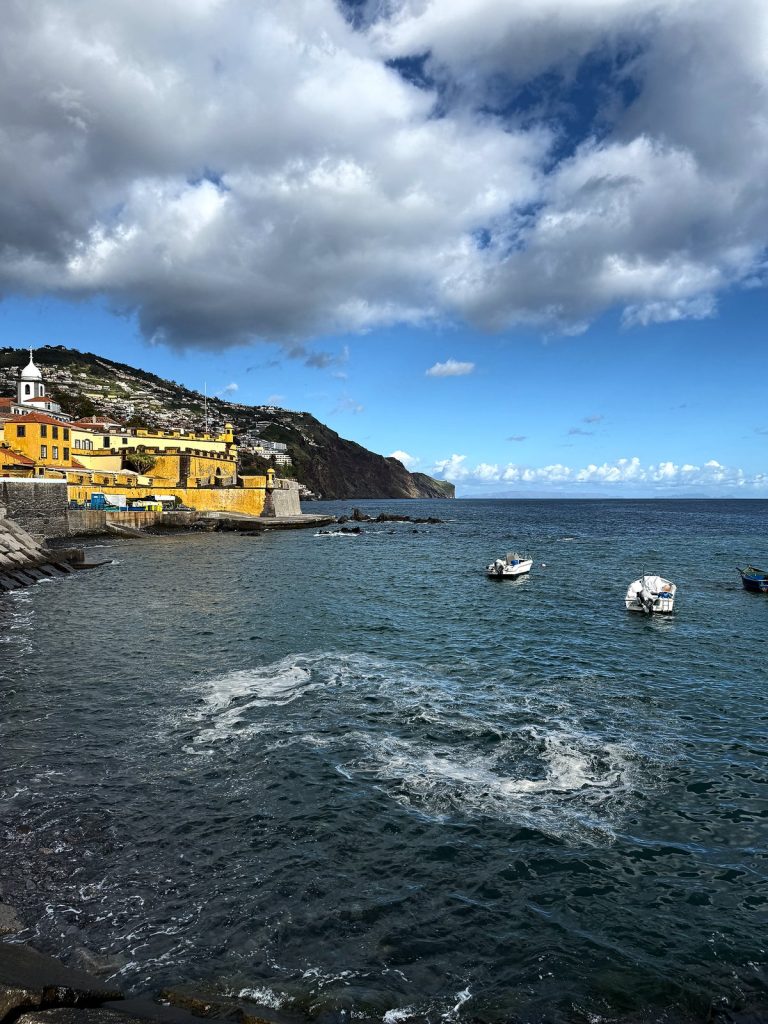 A bay with small boats, in the distance the yellow São Tiago fort, a white church tower and the hillside of houses in eastern Funchal