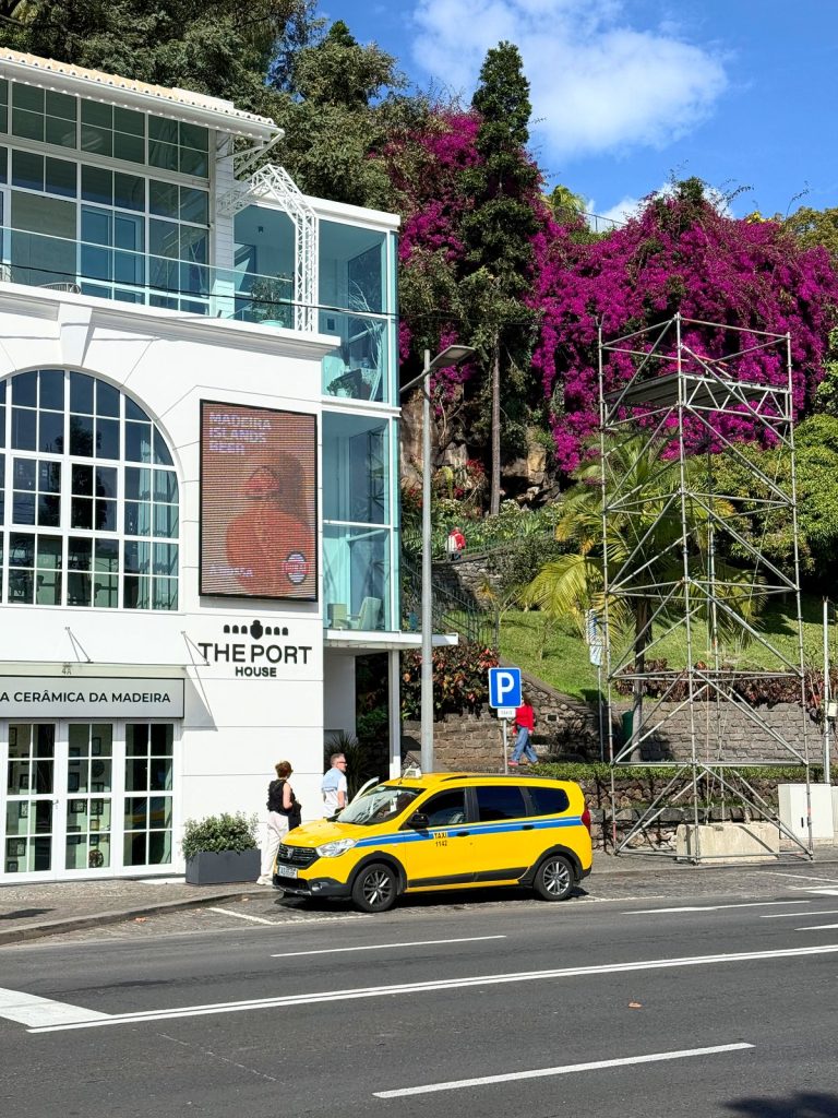 Yellow taxi in front of a white building with large windows, next to a flowering purple bougainvillea