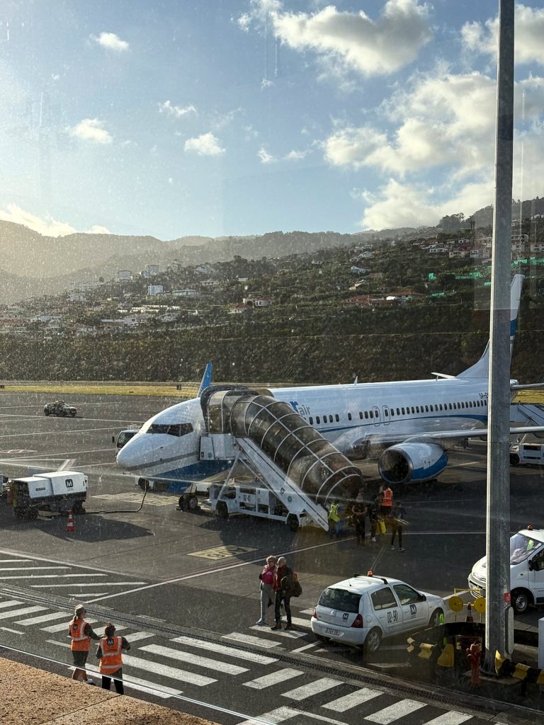 Passenger plane by the steps on the apron at Madeira airport, ground crew at work, a hillside with houses behind; view through the terminal window