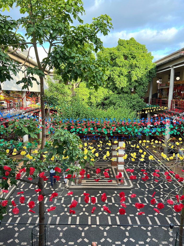 The courtyard of the Mercado dos Lavradores market hall with a mosaic floor, rows of colourful flower-shaped decorations overhead and galleries of stalls around it