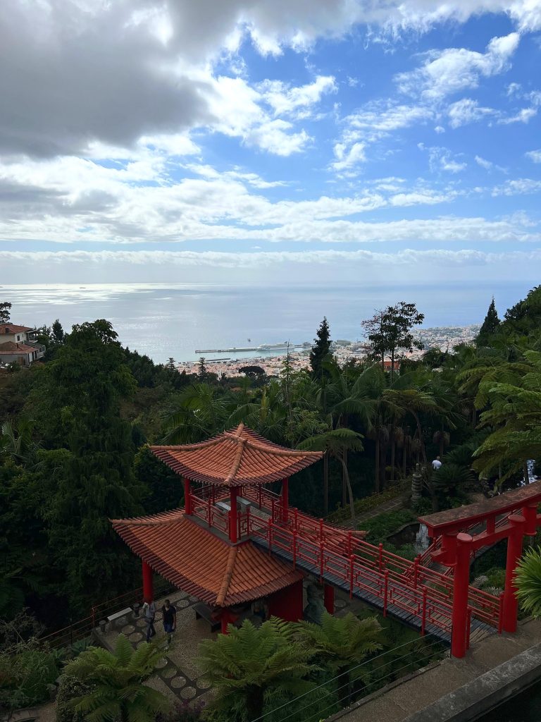 A red pagoda and bridge in the Oriental Garden at Monte Palace, with Funchal and the ocean under clouds in the distance