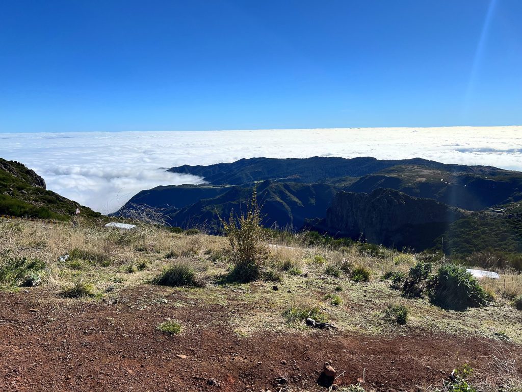 A sea of clouds below the mountain ridges near Pico do Arieiro, with red earth and dry shrubs in the foreground