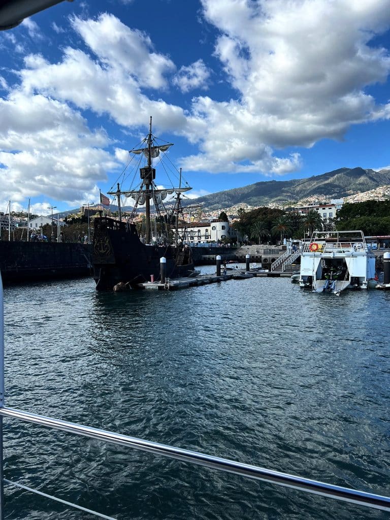 The wooden replica of the Santa Maria moored at the quay in Funchal harbour, with the city’s hillsides and clouds in a blue sky behind