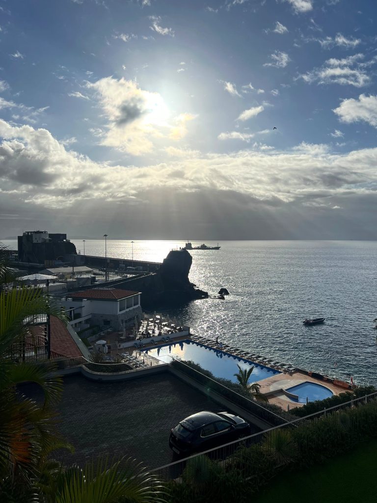 The sun breaking through clouds over the ocean, with hotel pools, a breakwater and a rock in the sea in western Funchal below