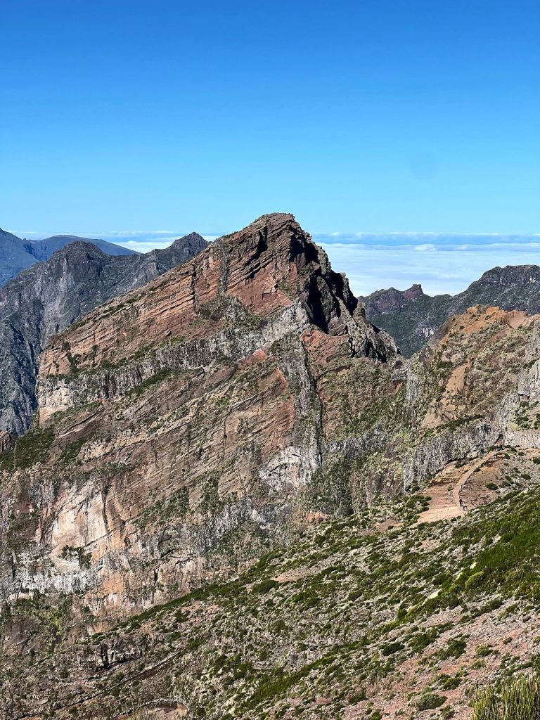 A reddish-brown rocky peak with layers of volcanic rock, other ridges and a sea of clouds behind