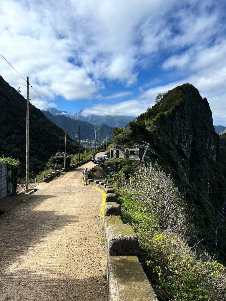 A narrow path along a stone wall above a steep green cliff, with mountains and clouds in the distance