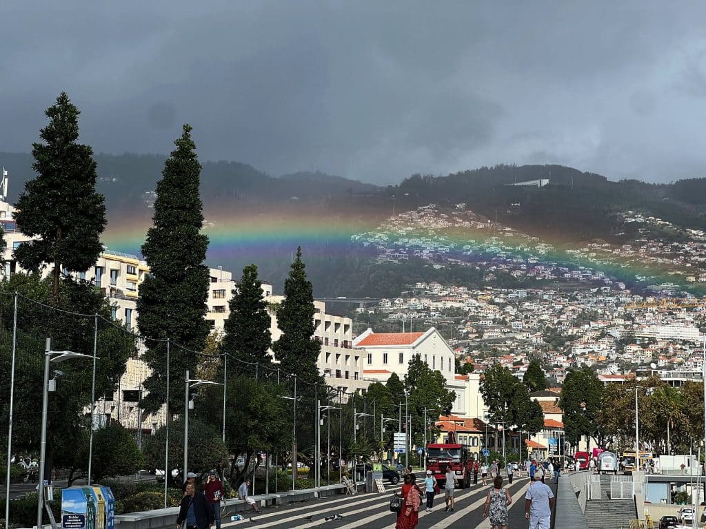 A rainbow over the hillsides of Funchal under dark clouds, with the tree-lined promenade, buildings and small figures of passers-by below