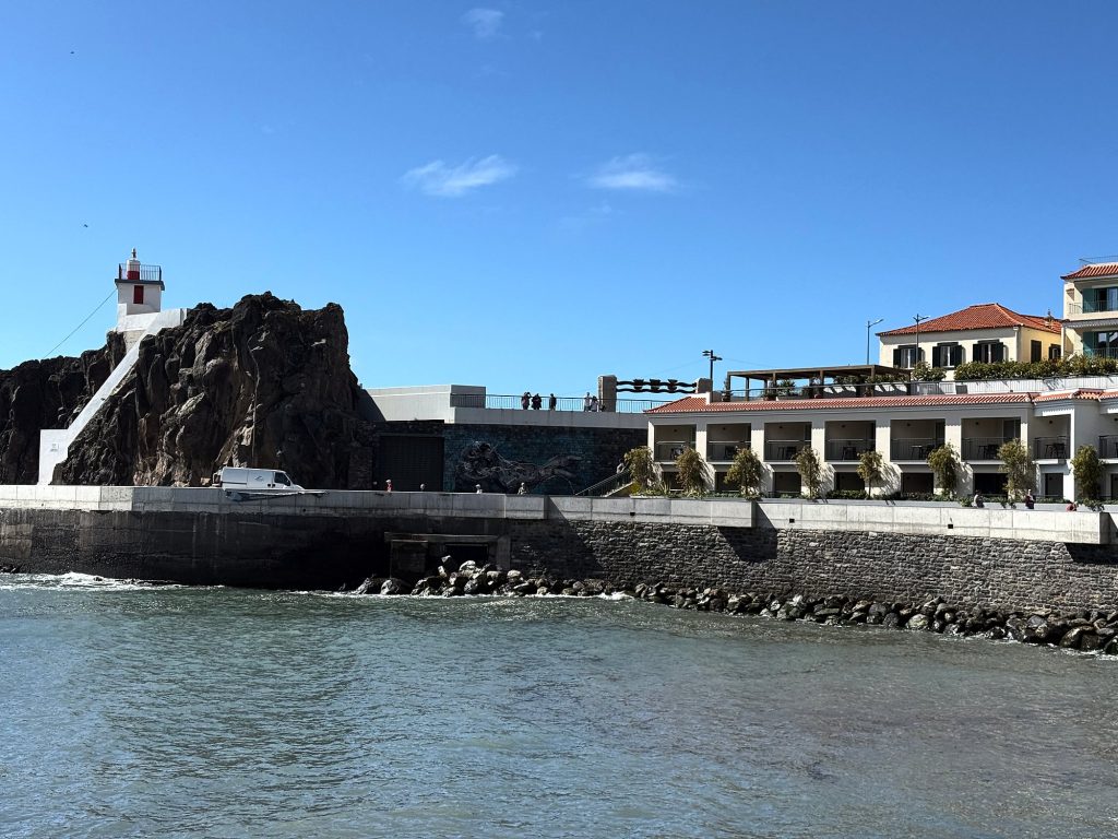 A small lighthouse on a black rock at the harbour entrance in Câmara de Lobos, next to the quay and a building with balconies