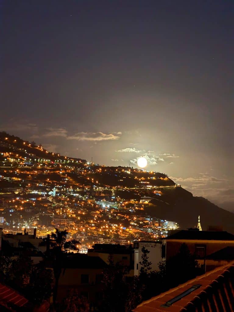 A full moon low over the lit-up hillside of Funchal at night, with rooftops in the foreground