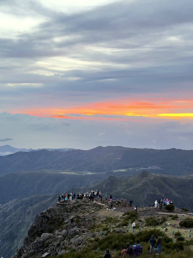 People on a rocky viewpoint at Pico do Arieiro at dawn, with an orange band of sky above the mountain ridges