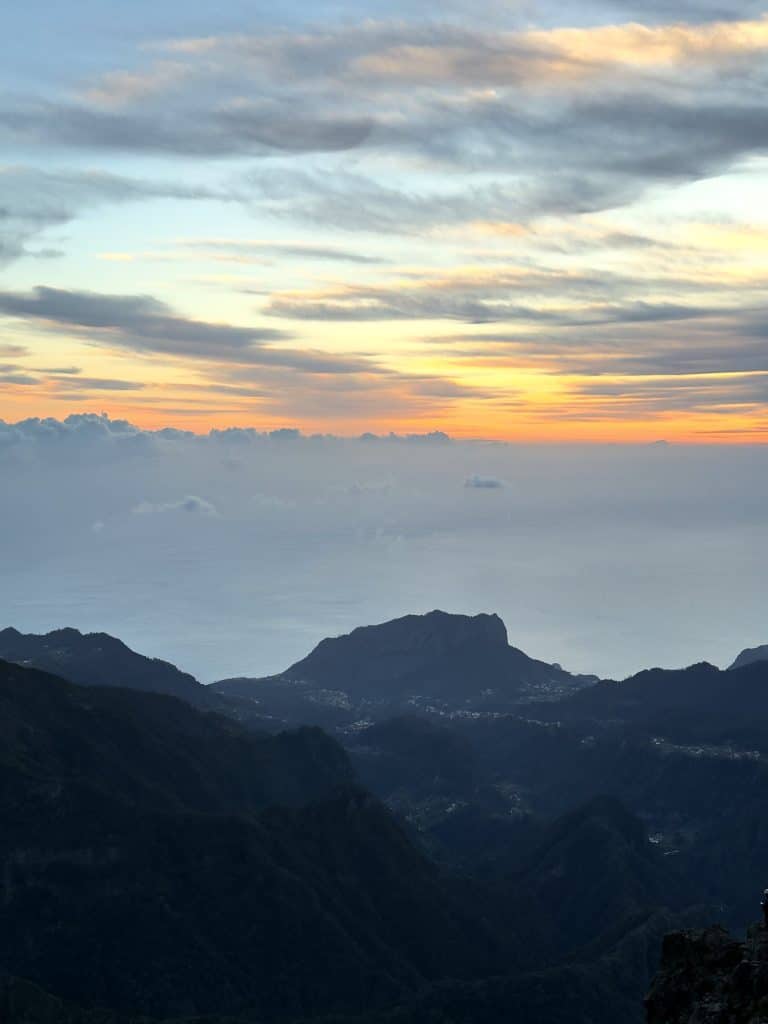 An orange sky at sunrise over a sea of clouds, with dark ridges and valleys below