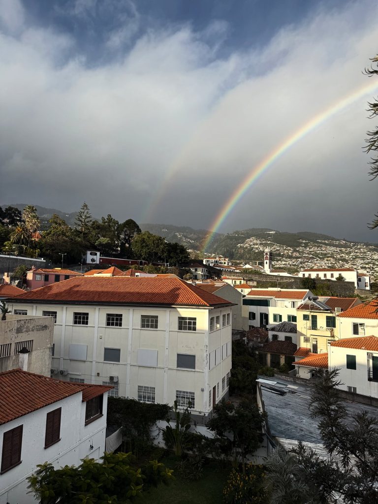 A double rainbow over the rooftops of Funchal under dark rain clouds, a white building with an orange roof in the foreground and houses on the slopes behind