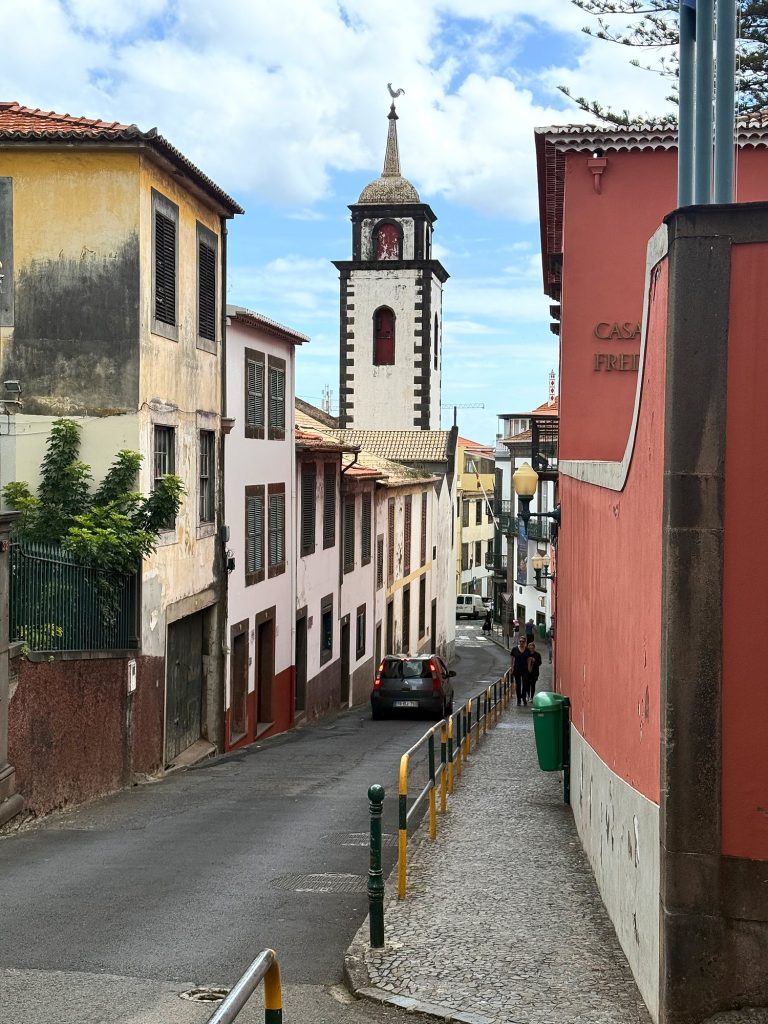 A narrow street with old town houses and a red house, a white church tower at the end