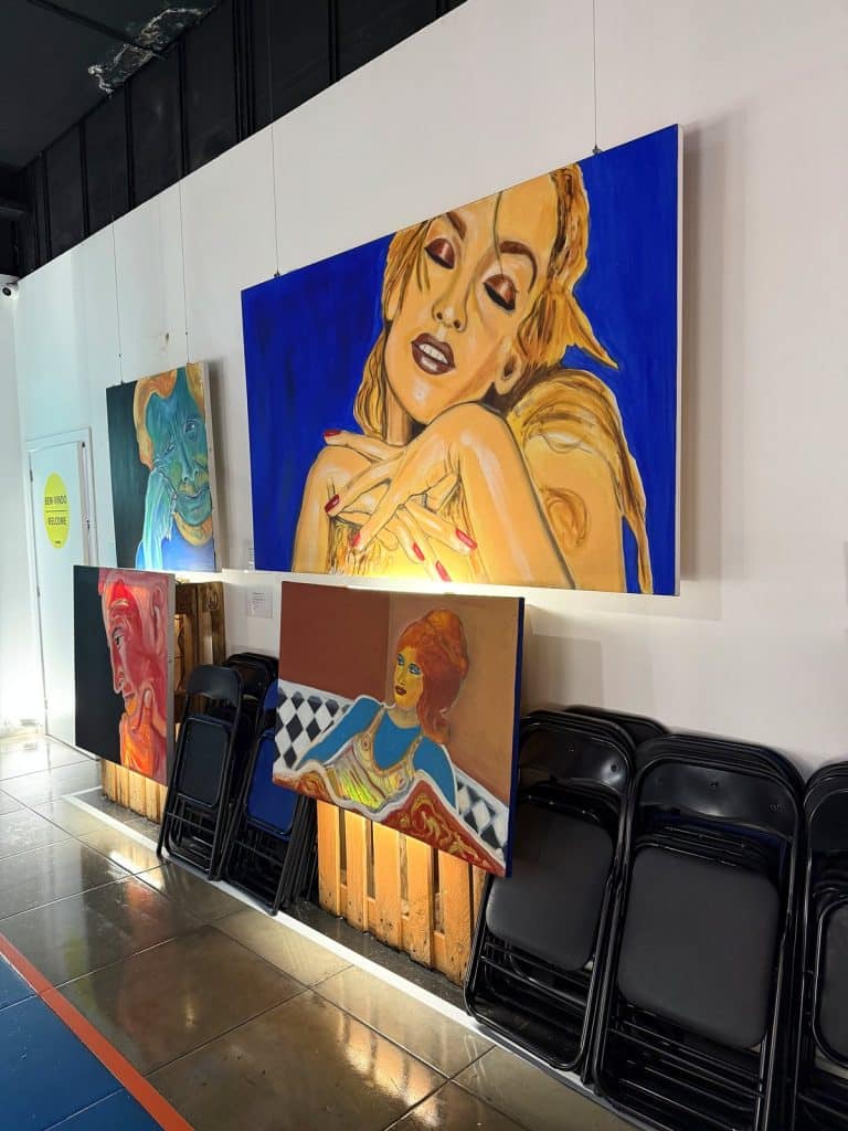 Large colourful pop-art paintings on the white wall of a corridor, with canvases leaning below them and folded chairs