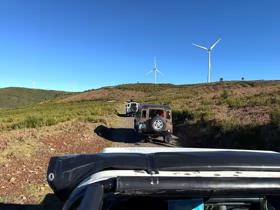 Land Rovers on a gravel road across the Paul da Serra plateau, seen from an open jeep, with wind turbines behind