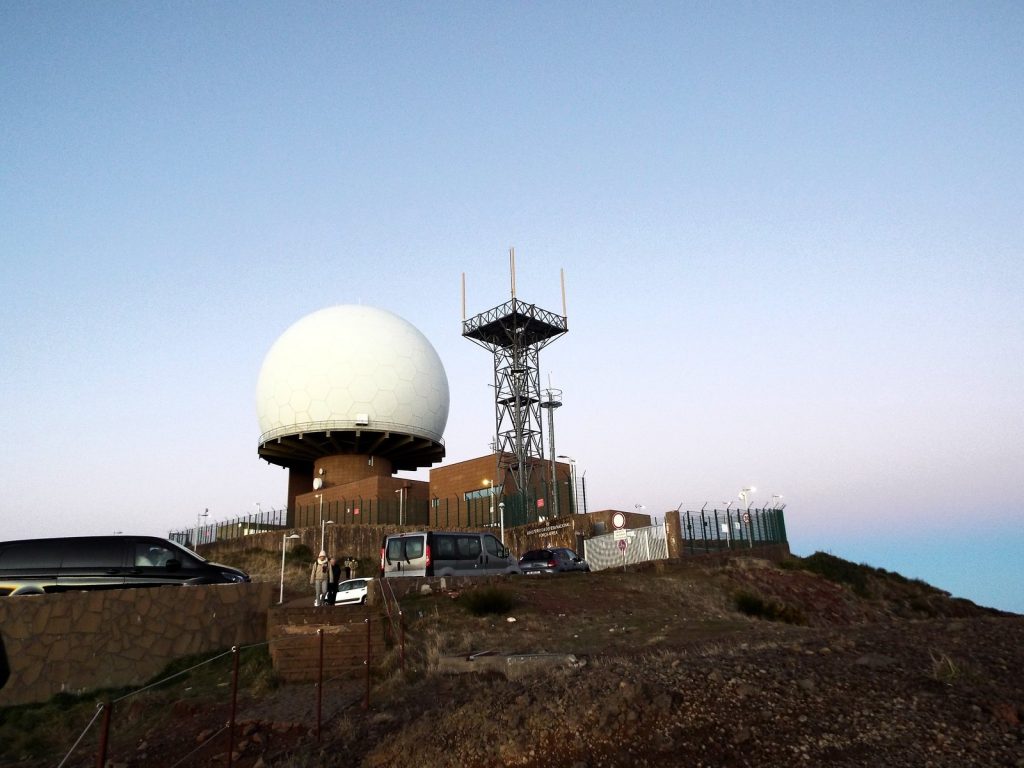 The radar dome and a steel mast with antennas on the summit of Pico do Arieiro, cars parked by the buildings, pink sky at dawn