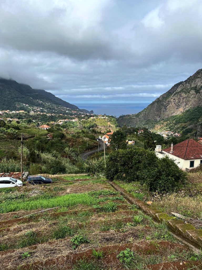 A wide green valley with fields and houses sloping down to the ocean between mountains in the clouds