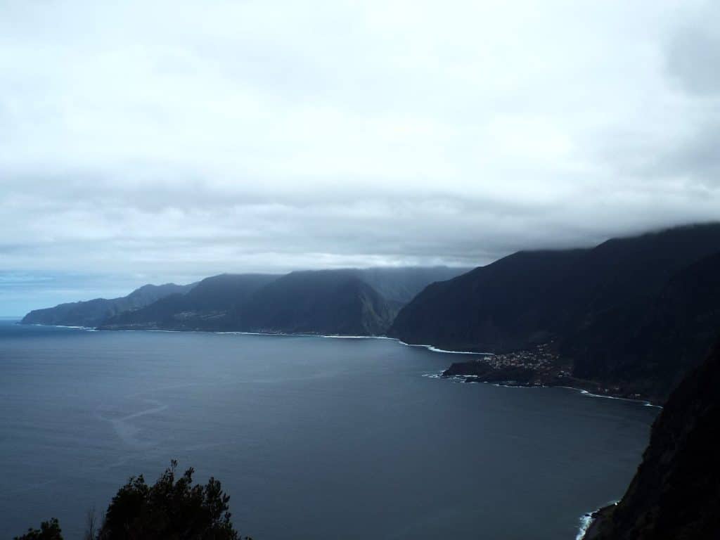 Overcast sky over Madeira’s north coast, dark cliffs dropping into a grey ocean, clouds hiding the peaks and a small village by the shore