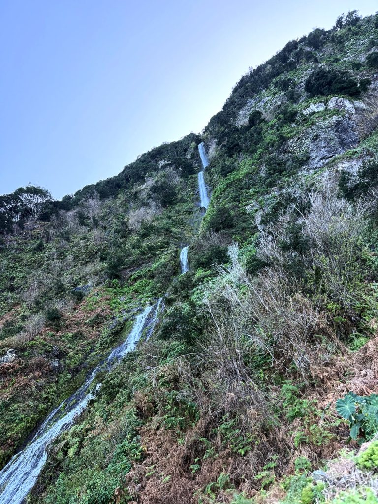 A narrow waterfall cascading down a steep, scrub-covered slope under a blue sky