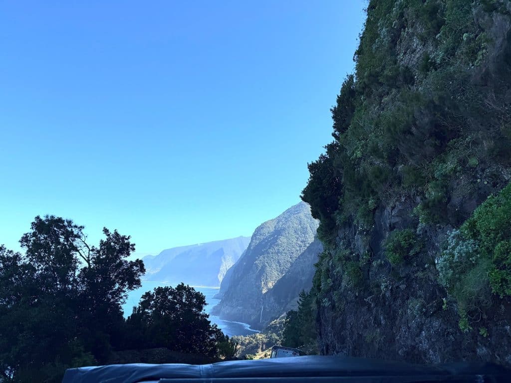 A wooded rock face above the road, with the north coast cliffs and the ocean in the distance and the roof of a jeep at the bottom of the frame
