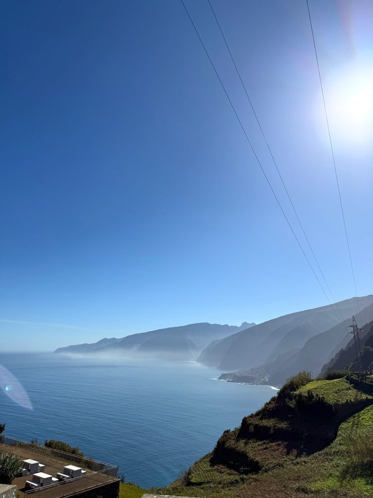 Madeira’s north coast from above: steep slopes dropping to the ocean, a town on a low headland, haze and sun, power lines on the right