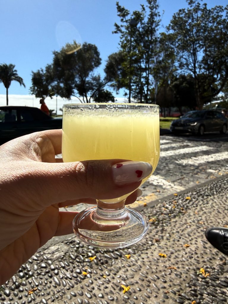 A hand holding a stemmed glass of pale yellow poncha, with a palm-lined street behind