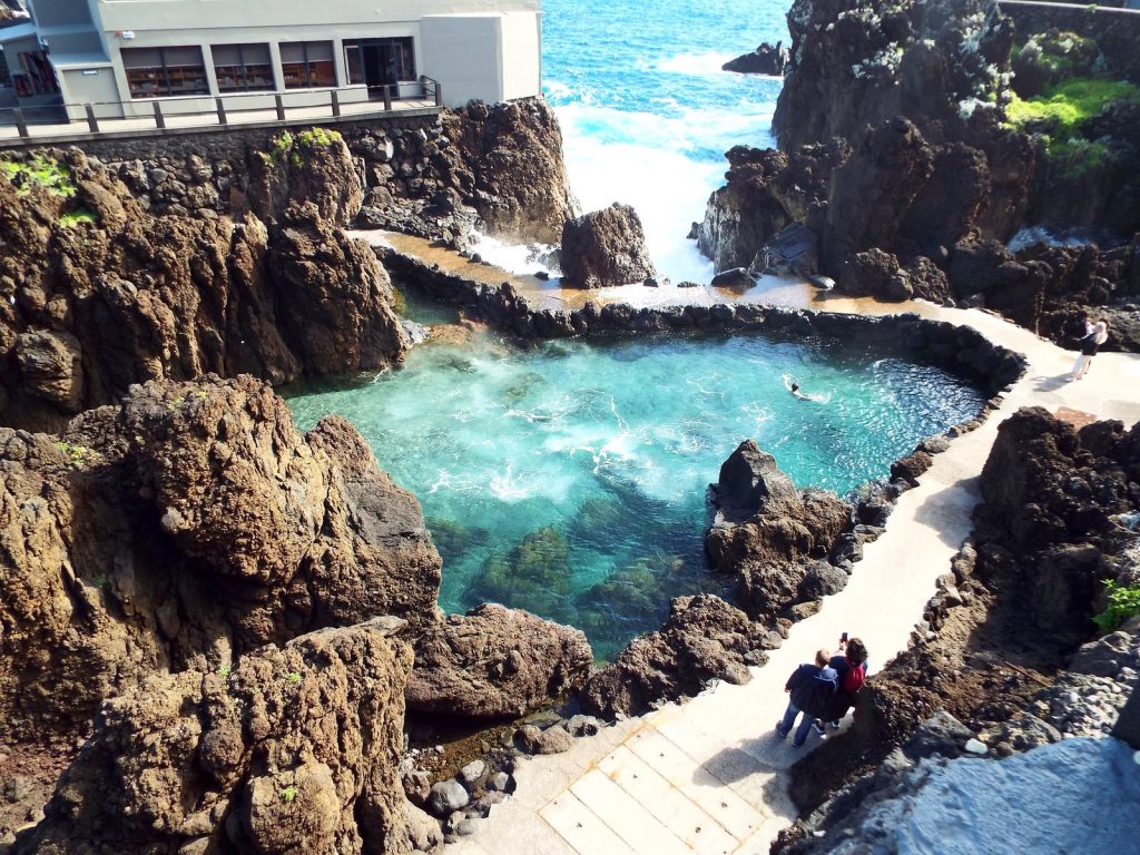 A natural pool with turquoise water among black volcanic rocks, a concrete path and a building on the shore, ocean waves beyond the rocks