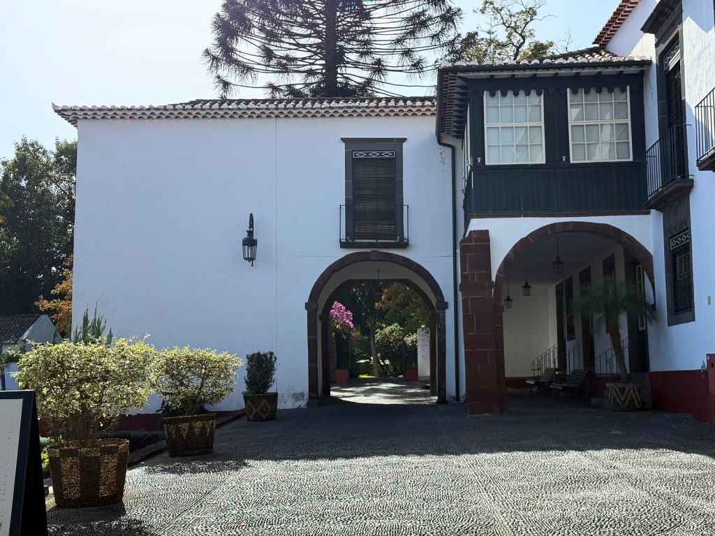A white manor house with dark wooden shutters and two arches, a cobbled courtyard with planters in front