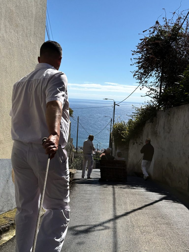Carreiro dressed in white steering a wicker toboggan down a steep street from Monte, with the ocean behind