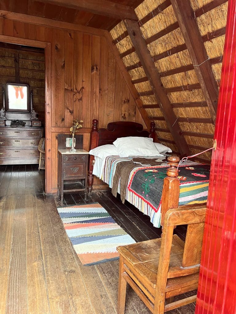 Inside a thatched house: a wooden bed with a colourful bedspread, a striped rug, a chair and a chest of drawers with a mirror